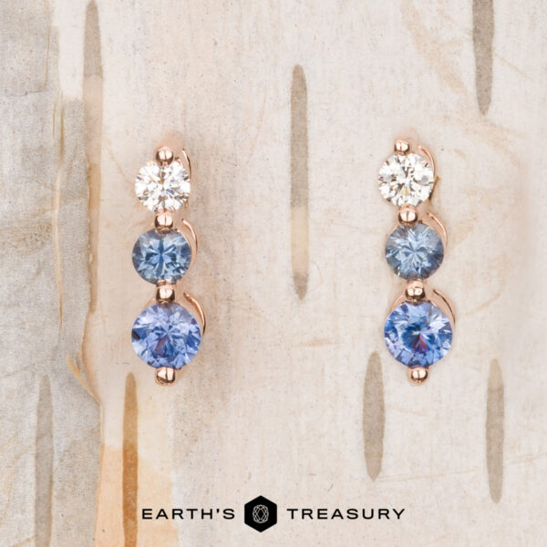 The "Dawn" Earrings in 14k rose gold Introducing The "Dawn" Earrings in 14k rose gold: a stunning trio of radiant white and blue gems elegantly arranged in a vertical design on a lightly textured background.