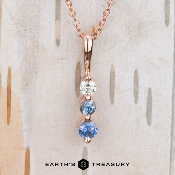The "Dawn" Necklace in 14k rose gold Introducing The "Dawn" Necklace in 14k rose gold, a breathtaking jewelry piece showcasing three gemstones in shades of white, light blue, and medium blue arranged vertically on a rose gold chain. Its elegance is exquisitely accentuated against a wooden background.