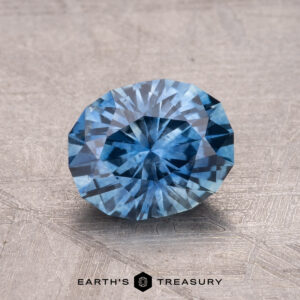 A 1.81-carat faceted round Montana sapphire, featuring a stunning blue hue, rests on a textured surface with the text "Earth's Treasury" below.
