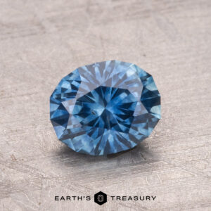 A faceted round blue 1.42-carat Montana sapphire (heated) is displayed on a textured surface, with the "Earth's Treasury" logo at the bottom.
