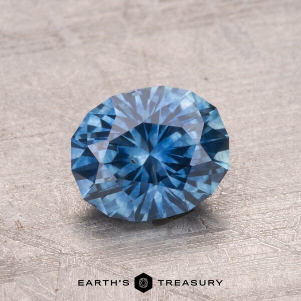 1.42-Carat Montana Sapphire (Heated) A faceted round blue 1.42-carat Montana sapphire (heated) is displayed on a textured surface, with the "Earth's Treasury" logo at the bottom.