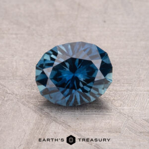 A brilliantly faceted round blue gem rests on a textured surface, with the "Earth's Treasury" logo elegantly displayed beneath it, showcasing the 1.28-Carat Montana Sapphire (Heated).