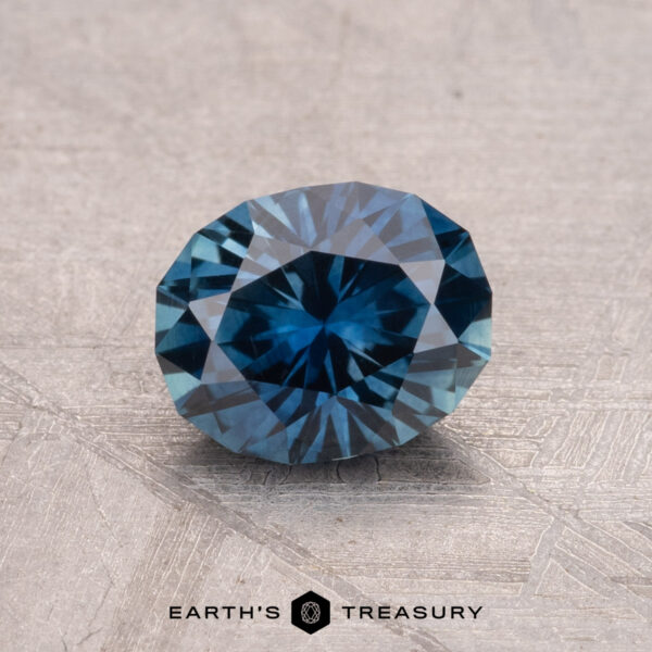1.28-Carat Montana Sapphire (Heated) A brilliantly faceted round blue gem rests on a textured surface, with the "Earth's Treasury" logo elegantly displayed beneath it, showcasing the 1.28-Carat Montana Sapphire (Heated).