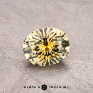 A 1.15-carat Montana Sapphire, featuring a round, brilliant cut with a lovely yellow hue, is elegantly displayed on a textured gray surface, accompanied by the "Earth's Treasury" logo beneath.