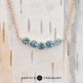 The "Breeze" Necklace in 14k yellow gold showcases a chain embellished with five round blue gemstones, beautifully set against a birch wood background.