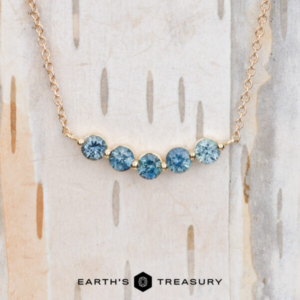 The "Breeze" Necklace in 14k yellow gold The "Breeze" Necklace in 14k yellow gold showcases a chain embellished with five round blue gemstones, beautifully set against a birch wood background.