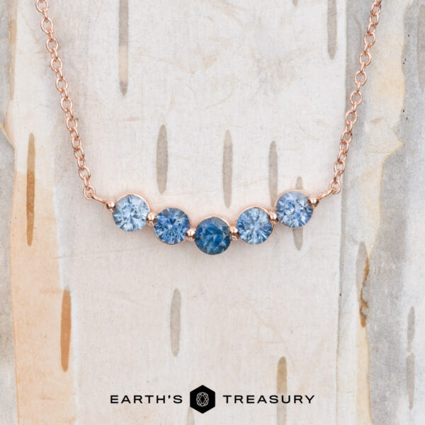 The "Breeze" Necklace in 14k rose gold The "Breeze" Necklace in 14k rose gold showcases five blue gemstones arranged in a row on a delicate chain, ideal for anyone who values elegant jewelry. Presented against a backdrop of rich wood texture, this piece exemplifies sophistication and style.