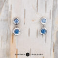 Two Dewdrop Earrings in 14k white gold with blue gemstones are displayed on a textured surface. The brand name "Earth's Treasury" is elegantly visible at the bottom.