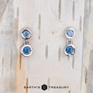 Two Dewdrop Earrings in 14k white gold with blue gemstones are displayed on a textured surface. The brand name "Earth's Treasury" is elegantly visible at the bottom.