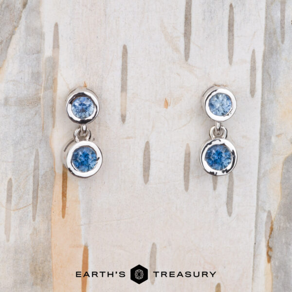 The "Dewdrop" Earrings in 14k white gold Two Dewdrop Earrings in 14k white gold with blue gemstones are displayed on a textured surface. The brand name "Earth's Treasury" is elegantly visible at the bottom.