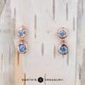 A pair of "Dewdrop" Earrings in 14k rose gold, featuring blue gemstones, is elegantly displayed on a textured wooden background.