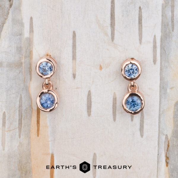 The "Dewdrop" Earrings in 14k rose gold A pair of "Dewdrop" Earrings in 14k rose gold, featuring blue gemstones, is elegantly displayed on a textured wooden background.