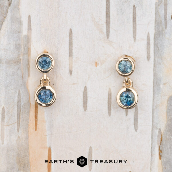 The "Dewdrop" Earrings in 14k yellow gold The "Dewdrop" Earrings in 14k Yellow Gold: Gleaming gold earrings embellished with radiant blue gemstones, gracefully displayed on a textured wooden background. The text at the bottom reads "Earth’s Treasury.