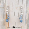 Pair of Journey Earrings in 14k white gold with blue and clear gemstones on a light wood background. Text at the bottom says "Earth's Treasury.