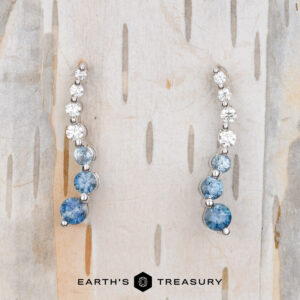 Pair of Journey Earrings in 14k white gold with blue and clear gemstones on a light wood background. Text at the bottom says "Earth's Treasury.