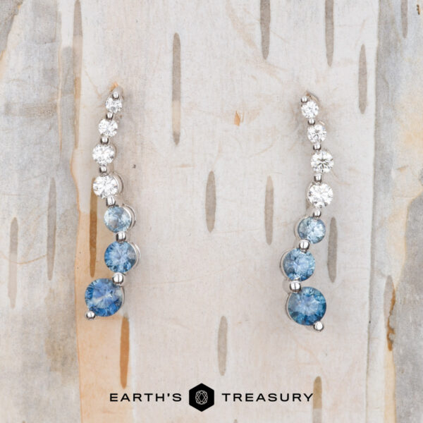 Pair of Journey Earrings in 14k white gold with blue and clear gemstones on a light wood background. Text at the bottom says "Earth's Treasury.