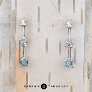The "Dusk" Earrings in 14k white gold, adorned with two blue gemstones and one clear gemstone on each earring, are elegantly displayed against a textured light wood backdrop. Beneath them, the "Earth's Treasury" logo subtly enhances the sophisticated presentation.