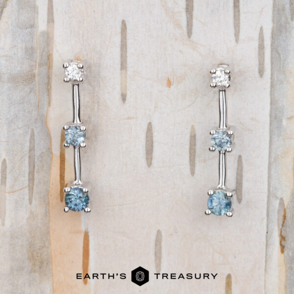 The "Dusk" Earrings in 14k white gold The "Dusk" Earrings in 14k white gold, adorned with two blue gemstones and one clear gemstone on each earring, are elegantly displayed against a textured light wood backdrop. Beneath them, the "Earth's Treasury" logo subtly enhances the sophisticated presentation.