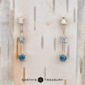 The "Dusk" Earrings in 14k yellow gold: Elegant dangle earrings featuring blue and clear gemstones, showcased on a light wooden background with vertical grooves.