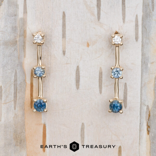 The "Dusk" Earrings in 14k yellow gold The "Dusk" Earrings in 14k yellow gold: Elegant dangle earrings featuring blue and clear gemstones, showcased on a light wooden background with vertical grooves.