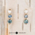 The "Dawn" Earrings in 14k yellow gold feature one round diamond and two shimmering blue gemstones, beautifully arranged against a light wood background.