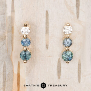 The "Dawn" Earrings in 14k yellow gold feature one round diamond and two shimmering blue gemstones, beautifully arranged against a light wood background.