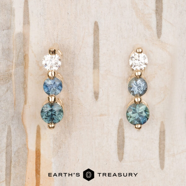 The "Dawn" Earrings in 14k yellow gold The "Dawn" Earrings in 14k yellow gold feature one round diamond and two shimmering blue gemstones, beautifully arranged against a light wood background.