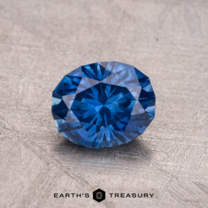 A 1.11-carat Montana Sapphire with a deep blue hue and intricate facets rests on a textured surface.