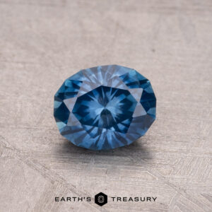 A sparkling 1.03-carat Montana sapphire, featuring a round faceted blue gemstone design, is placed on a textured gray surface.