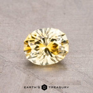 A 0.98-carat round, faceted yellow Montana sapphire rests on a textured gray surface with the "Earth's Treasury" logo at the bottom.