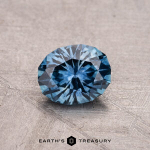 A round, faceted blue gemstone rests on a textured surface, labeled "0.90-Carat Montana Sapphire (Heated)" at the bottom.