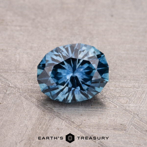 0.90-Carat Montana Sapphire (Heated) A round, faceted blue gemstone rests on a textured surface, labeled "0.90-Carat Montana Sapphire (Heated)" at the bottom.