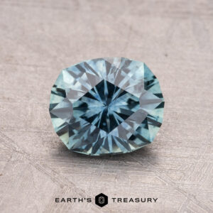 A faceted, round 1.33-carat Montana Sapphire with a blue-green hue is showcased against a textured gray surface. Text at the bottom reads "EARTH'S TREASURY.