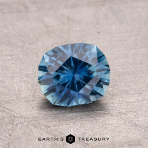 A 1.00-carat heated Montana Sapphire, exhibiting a faceted blue brilliance, rests on a textured surface alongside the "Earth's Treasury" logo.