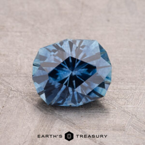 A 1.56-carat faceted blue Montana sapphire elegantly displayed on a light textured surface, with "Earth's Treasury" imprinted below.