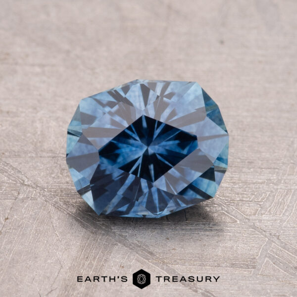 1.56-Carat Montana Sapphire (Heated) A 1.56-carat faceted blue Montana sapphire elegantly displayed on a light textured surface, with "Earth's Treasury" imprinted below.