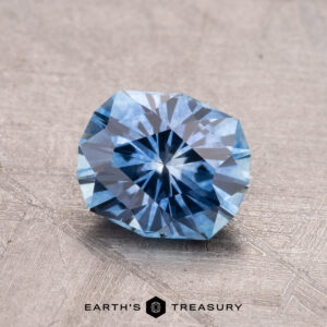 A faceted blue gemstone, named the 1.34-Carat Montana Sapphire (Heated), is displayed on a textured surface with the Earth’s Treasury logo visible at the bottom.