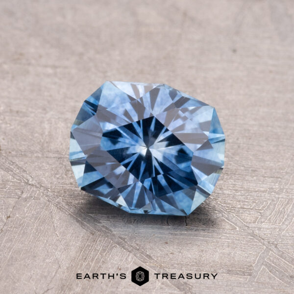 1.34-Carat Montana Sapphire (Heated) A faceted blue gemstone, named the 1.34-Carat Montana Sapphire (Heated), is displayed on a textured surface with the Earth’s Treasury logo visible at the bottom.