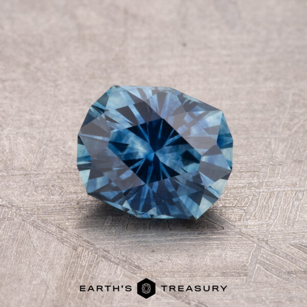 1.25-Carat Montana Sapphire (Heated) A round, faceted blue 1.25-carat Montana Sapphire (heated) on a textured surface with the text "Earth's Treasury" at the bottom.