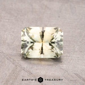 A 1.12-carat Montana sapphire in a faceted yellow-green hue is showcased on a textured surface, accompanied by the "Earth's Treasury" brand logo below.