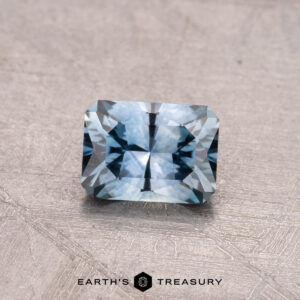 A 0.68-carat rectangular-cut blue Montana sapphire placed on a textured surface, with the text "Earth's Treasury" beneath it.