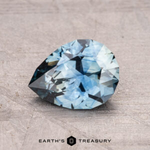 A pear-shaped blue gemstone, specifically a 1.41-carat Montana sapphire (heated), is displayed on a textured surface with the label "Earth's Treasury" at the bottom.
