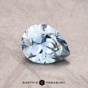 A pear-shaped 1.26-carat Montana sapphire, exhibiting facets and a blue hue, rests on a textured surface. The text "Earth's Treasury" is visible at the bottom.