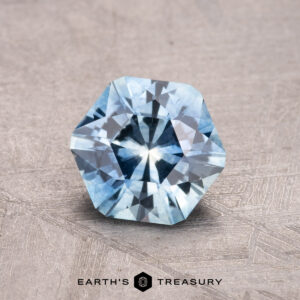 A 1.38-carat Montana sapphire (heated) with a star-shaped internal pattern is displayed against a textured gray surface.