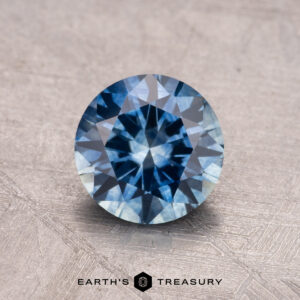 A 1.46-carat heated Montana Sapphire, cut in a round brilliant shape, sits on a textured gray surface with the "Earth's Treasury" logo at the bottom.