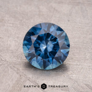 A round, faceted blue gemstone is displayed on a textured surface, with "1.43-Carat Montana Sapphire (Heated)" written below.