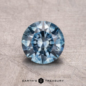 A 1.24-carat Montana sapphire, heated, in a round cut with intricate facets rests on a textured surface. The text "Earth's Treasury" appears at the bottom.