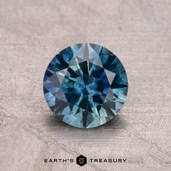 1.19-Carat Montana Sapphire (Heated) A round blue 1.19-carat Montana sapphire, known for its intricate facets, is displayed on a textured surface, with the "Earth's Treasury" text and logo below.