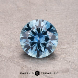 A round blue Montana sapphire, weighing 1.08 carats and featuring intricate facets, rests on a textured surface with "Earth's Treasury" printed below.
