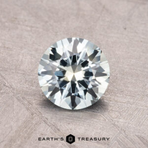 A faceted, round gemstone with a light blue hue, displayed on a textured surface. Near the bottom, the text reads "1.05-Carat Montana Sapphire (Heated) by Earth's Treasury.
